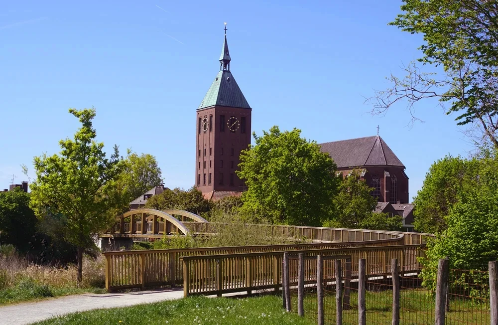 Blick auf die katholische Pfarrkirche St. Cyriakus in Weeze an einem sonnigen Frühlingstag. Im Vordergrund verläuft ein geschwungener Holzsteg durch eine grüne Parklandschaft mit Wiesen, Bäumen und Sträuchern. Dahinter erhebt sich der markante Backsteinturm der Kirche mit seiner spitzen Turmhaube und den sichtbaren Zifferblättern der Turmuhr. Der klare blaue Himmel und das frische Grün der Bäume verleihen der Szene eine ruhige und freundliche Atmosphäre.