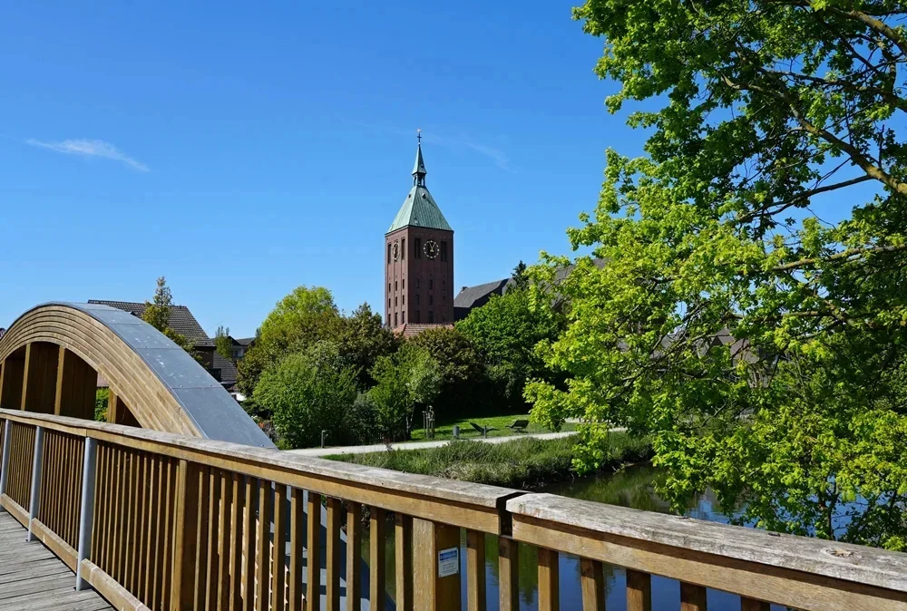 Blick von einer modernen Holzbrücke über die Niers auf die katholische Pfarrkirche St. Cyriakus in Weeze. Im Vordergrund prägt das hölzerne Brückengeländer das Bild, links ist der geschwungene Bogen der Brückenkonstruktion zu sehen. Dahinter spiegeln sich Bäume und Sträucher im ruhigen Wasser. Über dem frischen Frühlingsgrün erhebt sich der markante Backsteinturm der Kirche mit seiner spitzen Turmhaube. Ein strahlend blauer Himmel und das leuchtende Laub eines großen Baumes auf der rechten Bildseite sorgen für eine freundliche und idyllische Niederrhein-Stimmung.