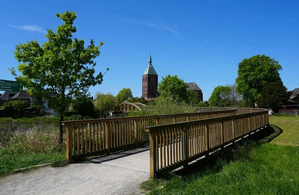 Geschwungener Holzsteg in einer grünen Parklandschaft in Weeze. Der Weg führt über die Niers und lenkt den Blick auf die katholische Pfarrkirche St. Cyriakus im Hintergrund. Der markante Backsteinturm mit seiner spitzen Turmhaube ragt zwischen Bäumen und Sträuchern hervor. Eine zweite Bogenbrücke ist in der Ferne zu erkennen. Die Szene wird von saftigem Frühlingsgrün, einer großen Baumkrone und einem klaren blauen Himmel geprägt und vermittelt eine ruhige, naturnahe Atmosphäre.