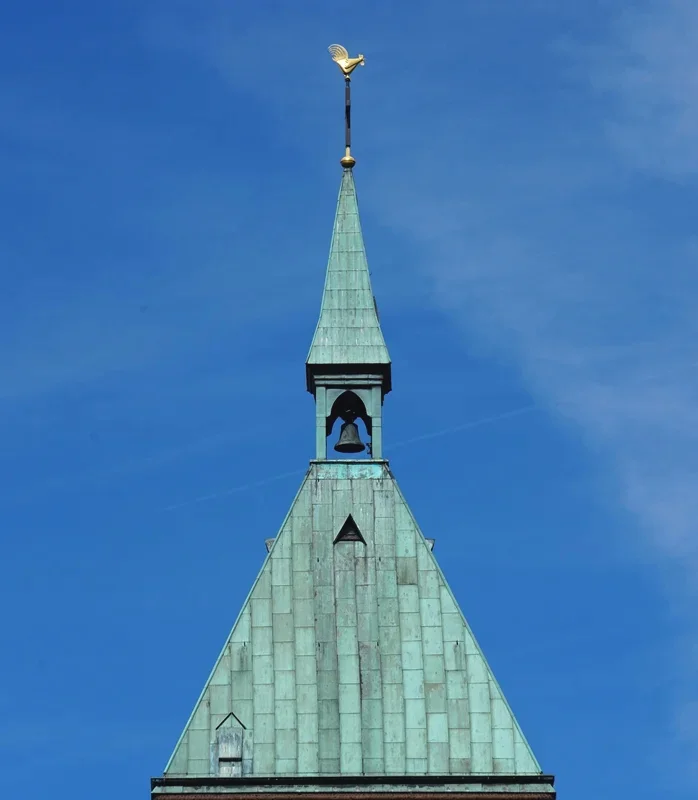 Detailaufnahme der Kirchturmspitze von St. Cyriakus in Weeze. Das kupfergrüne Turmdach mit offenem Glockenstuhl hebt sich vor einem tiefblauen Himmel ab. An der Spitze sitzt ein goldener Wetterhahn, der weithin sichtbar über dem historischen Kirchturm thront.