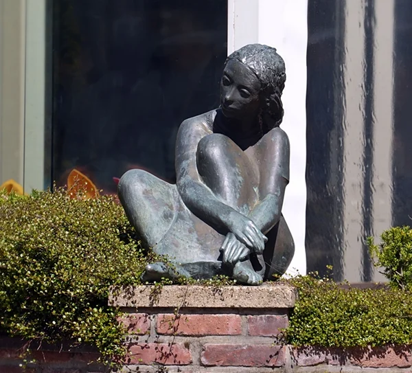 Bronzefigur einer sitzenden Frau vor dem Marktcafé in Weeze. Die Skulptur zeigt eine nachdenklich wirkende Person mit angezogenen Beinen und verschränkten Armen, die auf einem niedrigen Backsteinsockel sitzt. Umgeben wird die Figur von gepflegten Buchsbaumhecken und Zierpflanzen. Im Hintergrund sind die Fenster und Fassadenelemente des Gebäudes zu erkennen. Das Sonnenlicht hebt die Struktur und Patina der Bronze hervor und verleiht der Szene eine ruhige und beschauliche Atmosphäre im Ortskern von Weeze.