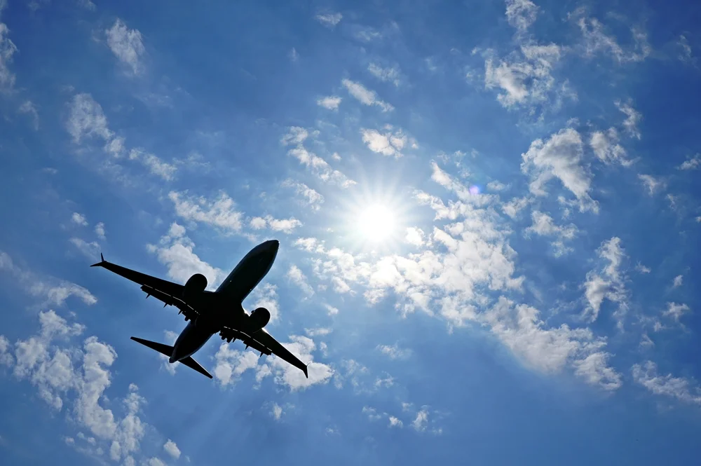 Ein Passagierflugzeug im Landeanflug auf den Weezer Flughafen fliegt als dunkle Silhouette vor einem strahlend blauen Himmel. Die Sonne steht hoch zwischen lockeren weißen Wolken und erzeugt helle Lichtstrahlen sowie einen leichten Lensflare-Effekt. Die Aufnahme entstand aus einer ungewöhnlichen Perspektive direkt von unten und vermittelt Dynamik und Sommerstimmung.
