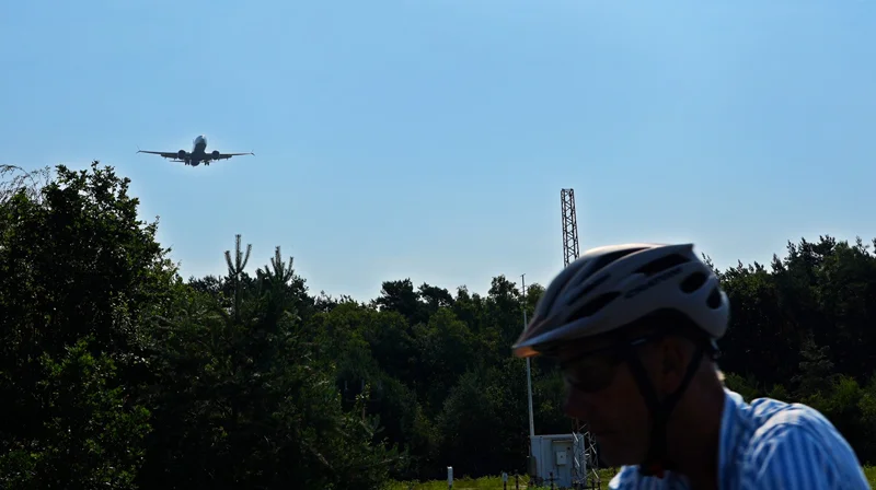 Radfahrer mit Fahrradhelm am Flughafen Weeze, während im Hintergrund ein Passagierflugzeug im Landeanflug über die Baumkronen fliegt.