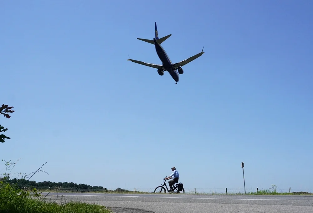 Blick auf einen Radfahrer vor der Start- und Landebahn des Flughafens Weeze. Über ihm fliegt ein Passagierflugzeug im Landeanflug durch den blauen Sommerhimmel.