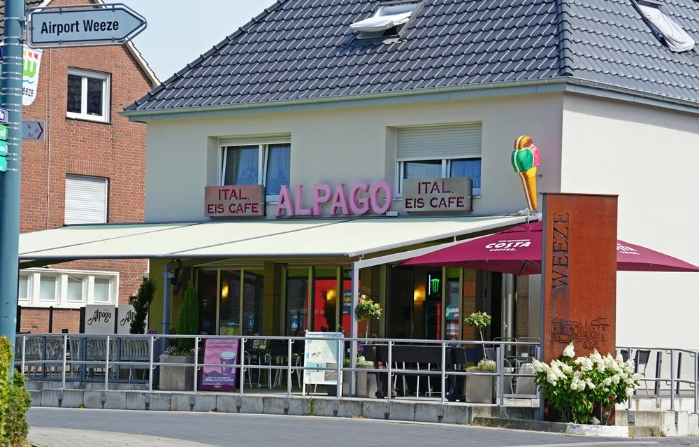 Eiscafé Alpago im Ortszentrum von Weeze mit großer Außenterrasse und markantem Schriftzug über dem Eingang. Links weist ein Straßenschild zum Airport Weeze, rechts steht eine Stele mit der Aufschrift „Weeze“.