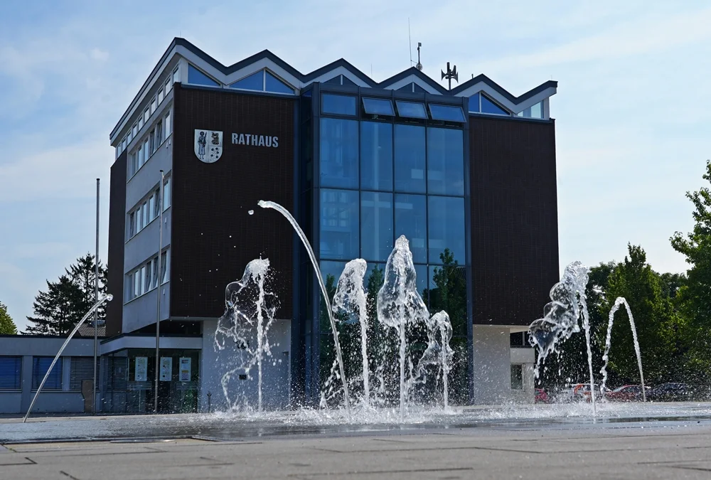 Modernes Rathaus der Gemeinde Weeze mit markanter Glasfassade und gezackter Dachform. Im Vordergrund sorgt ein Springbrunnen mit mehreren Wasserfontänen für einen lebendigen Blickfang.