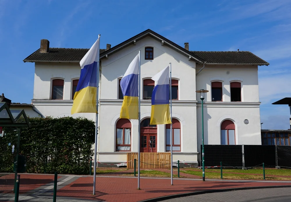 Frontalansicht des historischen Bahnhofs Weeze mit weißer Fassade und drei Fahnen vor dem Eingangsbereich.