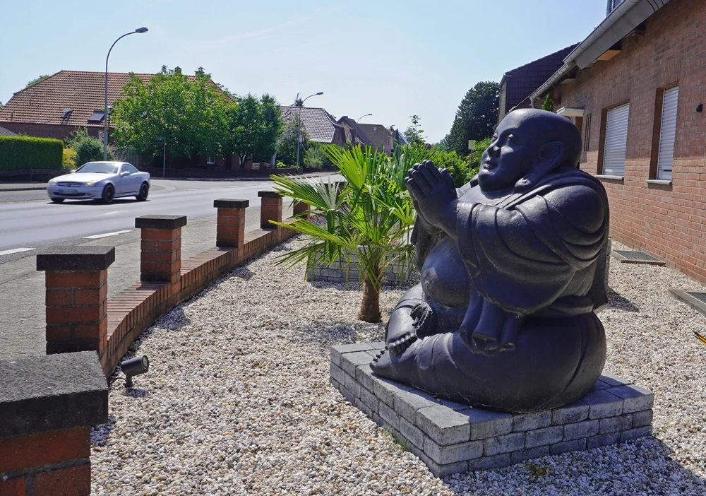 Ein großer sitzender Buddha in einem Vorgarten am Ortseingang von Wemb. Im Hintergrund sind Wohnhäuser und eine Straße zu sehen.