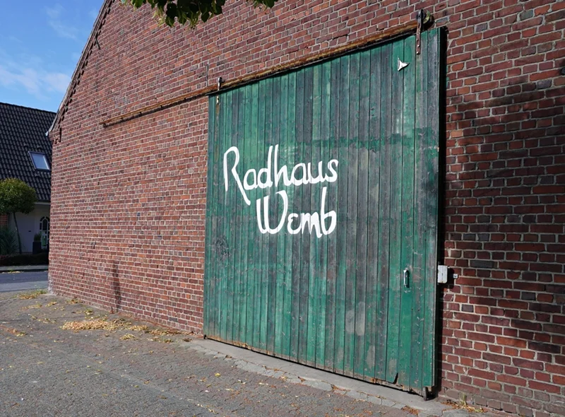 Grüne Scheunentür mit dem handgemalten Schriftzug „Radhaus Wemb“ an einem Backsteingebäude in Wemb.