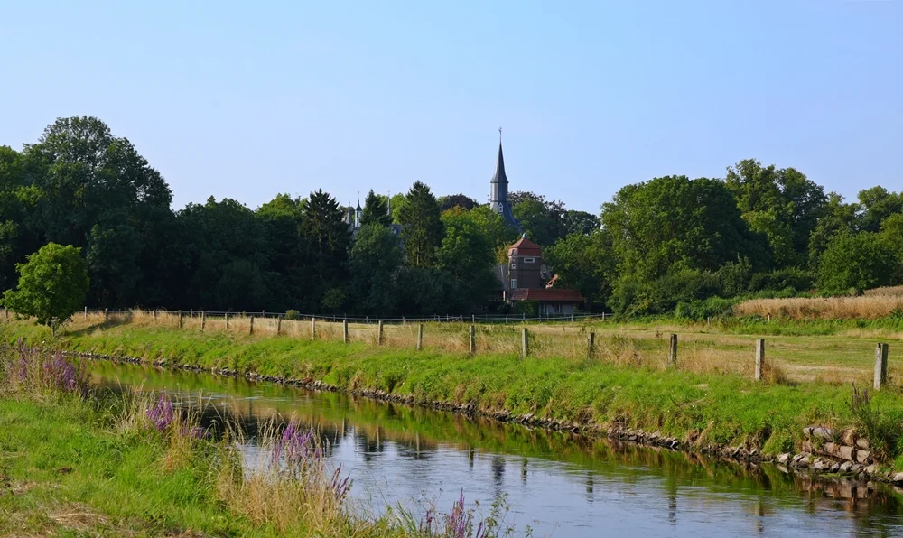 Blick über die Niers auf Wiesen und Bäume mit dem zwischen den Bäumen sichtbaren Turm von Schloss Wissen in Weeze