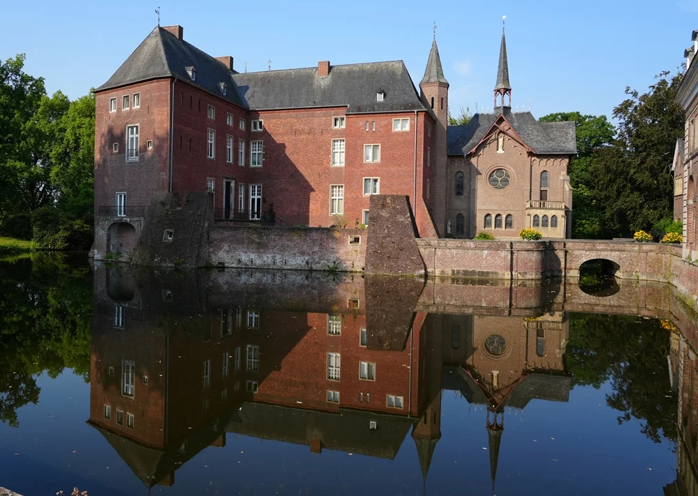 Blick auf Schloss Wissen in Weeze mit den historischen Gebäuden, der Kapelle und einer klaren Spiegelung im ruhigen Wassergraben.
