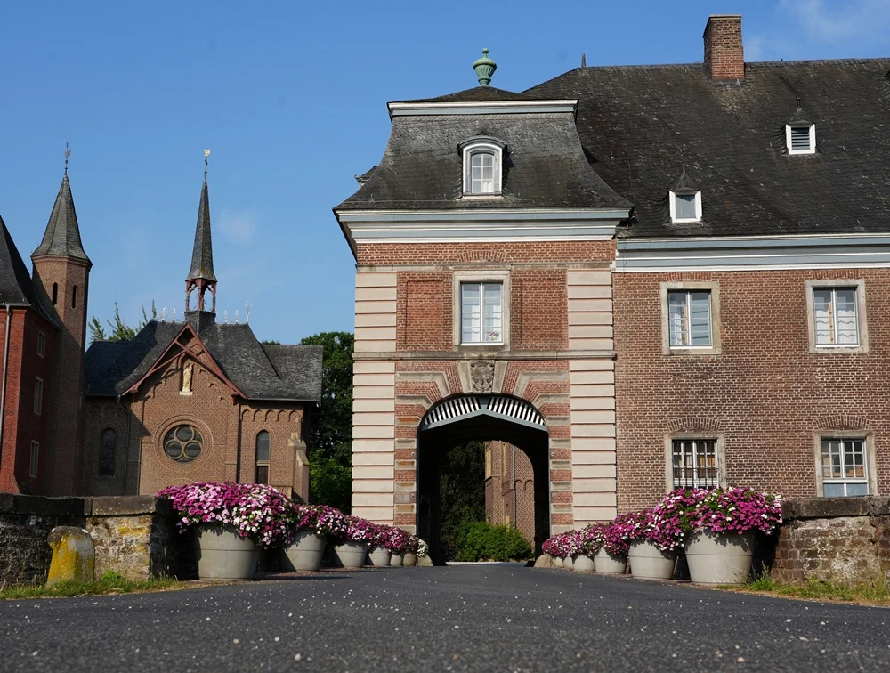 Blick auf das Eingangstor von Schloss Wissen in Weeze mit den Blumenkübeln entlang der Brücke und der Kapelle im Hintergrund.