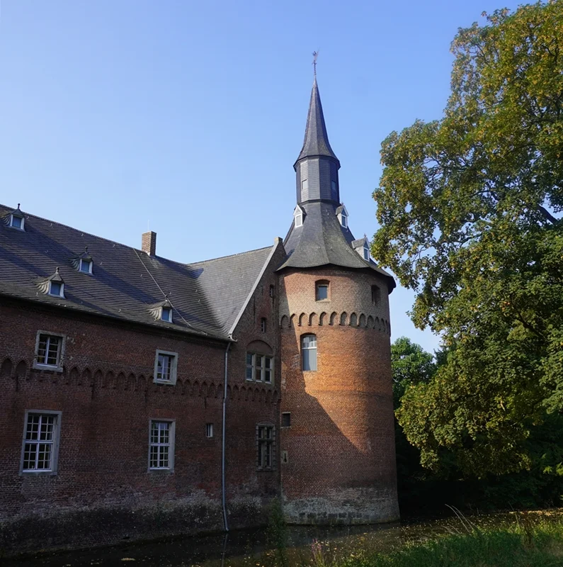 Der Eckturm von Schloss Wissen in Weeze mit historischem Backsteinmauerwerk, Wassergraben und altem Baumbestand.