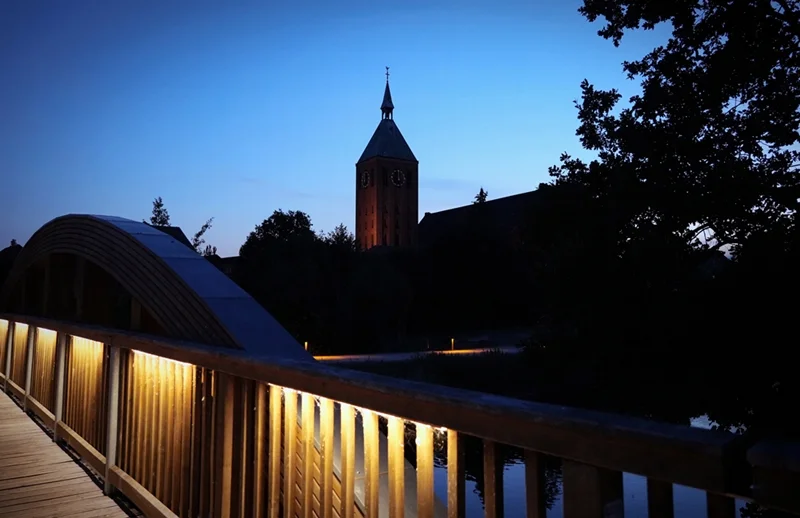 Beleuchtete Holzbrücke an der Nierspromenade in Weeze mit Kirchturm in der Abenddämmerung