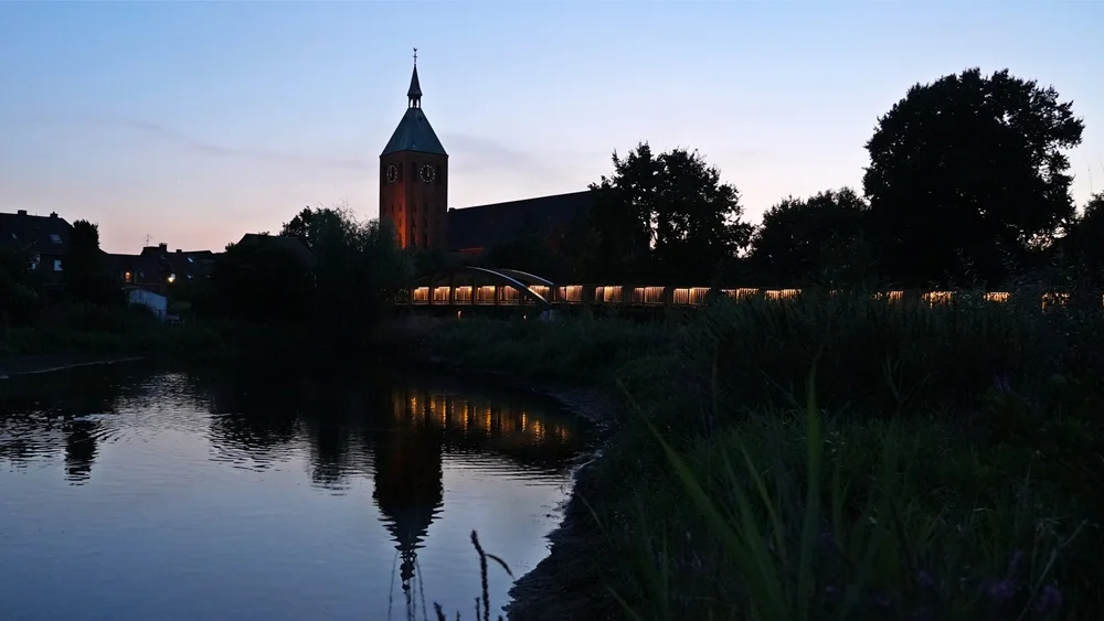 Kirchturm und beleuchtete Holzbrücke an der Niers in Weeze bei Abenddämmerung mit Spiegelung im Wasser