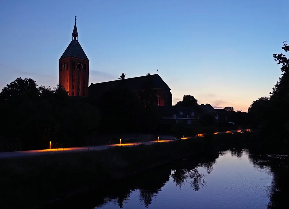 Beleuchtete Kirche an der Nierspromenade in Weeze bei Abenddämmerung mit Spiegelung in der Niers