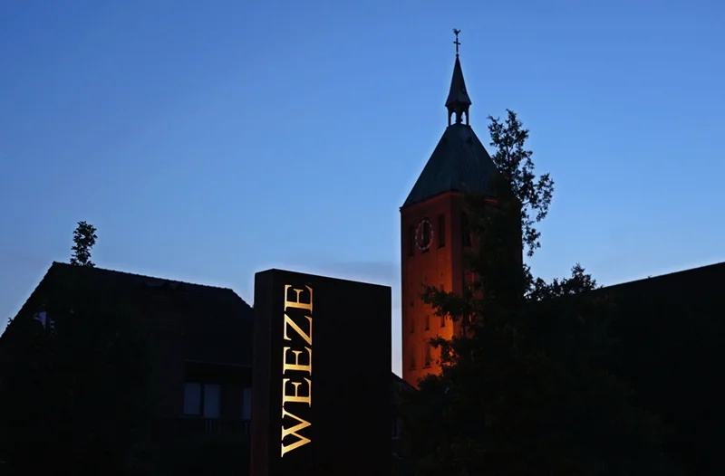 Beleuchteter Kirchturm in Weeze bei Abenddämmerung mit dem beleuchteten WEEZE-Schriftzug im Vordergrund