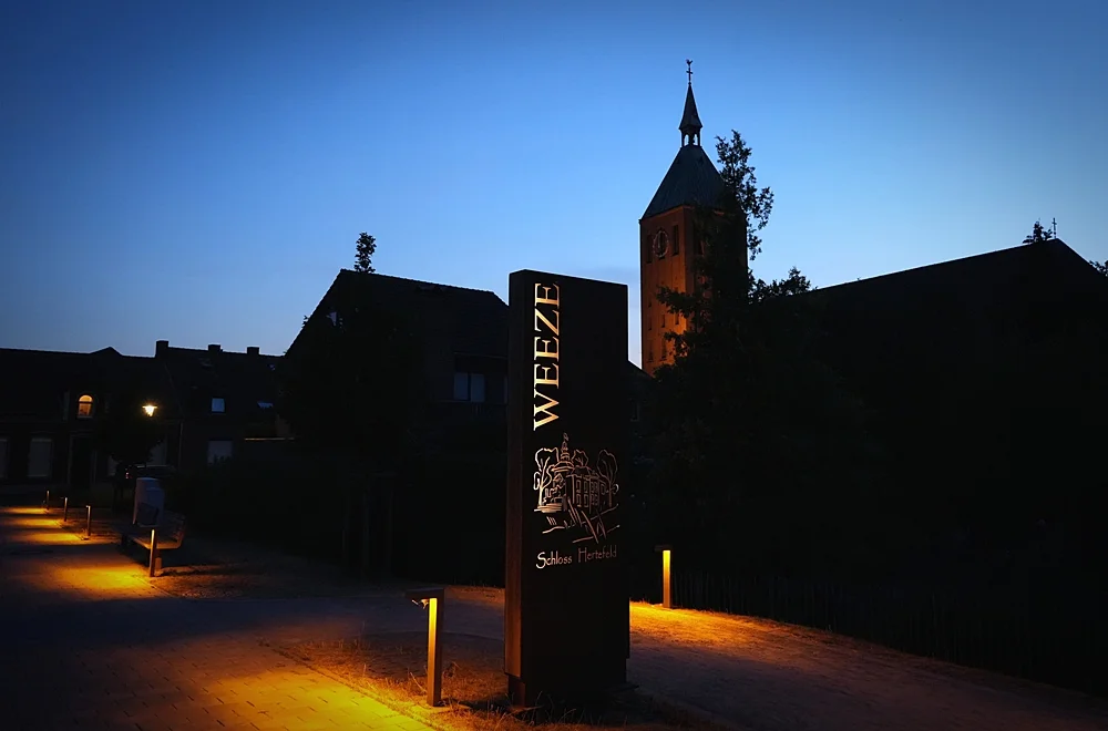 Beleuchteter WEEZE-Schriftzug am Schloss Hertefeld mit Kirchturm in der Abenddämmerung
