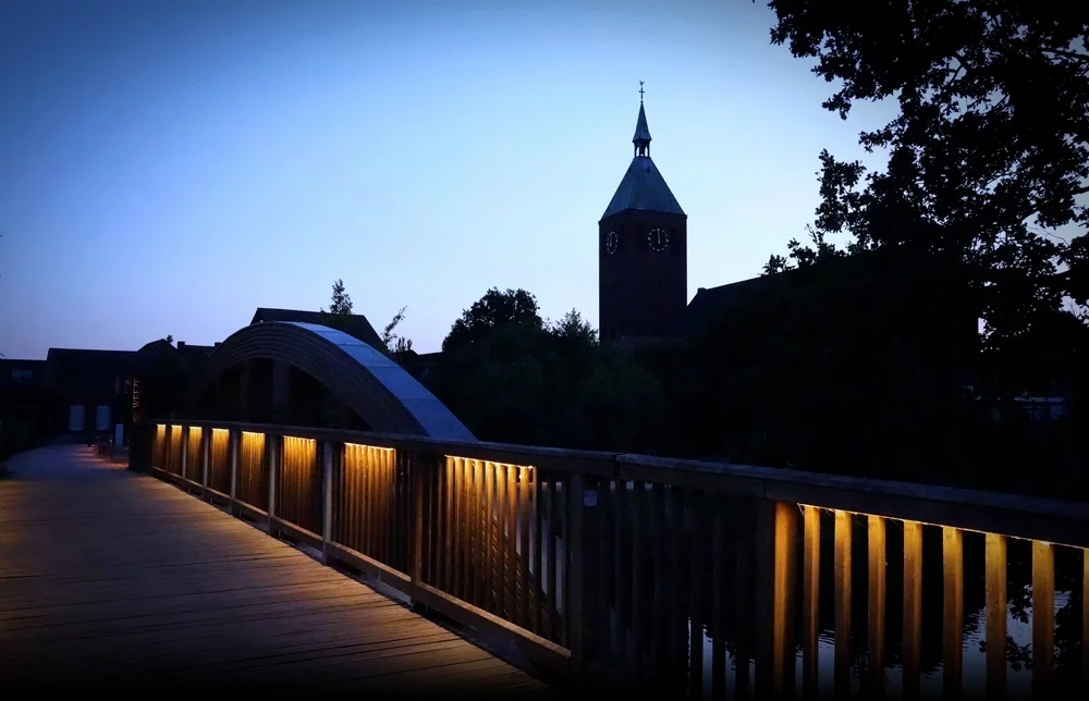 Beleuchtete Holzbrücke an der Nierspromenade in Weeze mit Kirchturm in der blauen Abenddämmerung