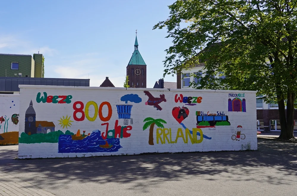Bunt bemalte Mauer zum Thema 800 Jahre Weeze mit dem Kirchturm von St. Cyriakus im Hintergrund