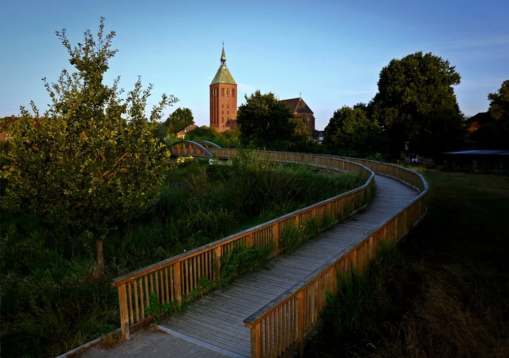 Blick über den geschwungenen Holzsteg auf die Kirche St. Cyriakus in Weeze am frühen Morgen