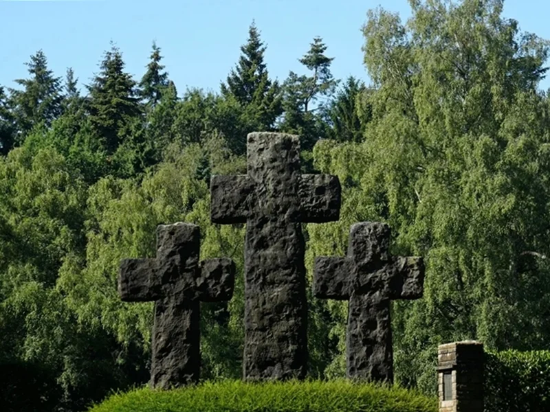 Drei große Steinkreuze des Mahnmals auf der Kriegsgräberstätte in Weeze vor grünen Bäumen