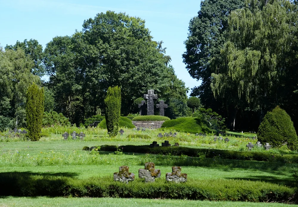 Weite Ansicht der Kriegsgräberstätte in Weeze mit Wildblumen, Steinkreuzen und altem Baumbestand