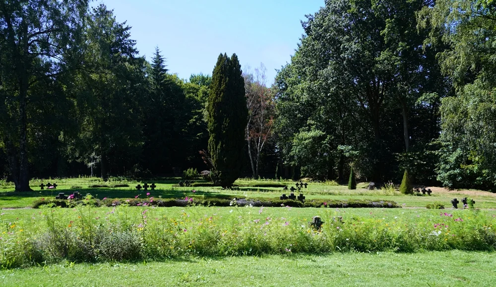 Weite Ansicht der Kriegsgräberstätte in Weeze mit Wildblumen, Steinkreuzen und altem Baumbestand