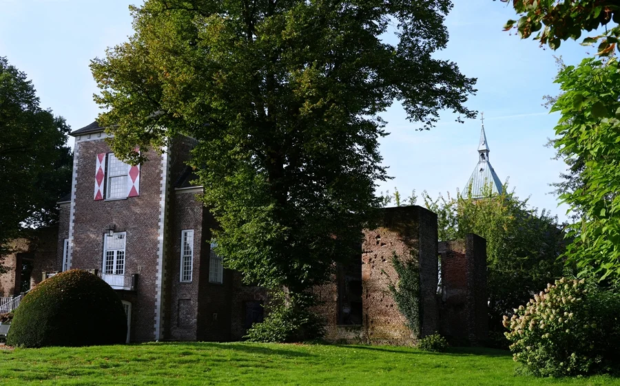 Seitenansicht von Schloss Hertefeld in Weeze mit historischem Mauerwerk, großer Parkanlage und dem Kirchturm von St. Cyriakus im Hintergrund.