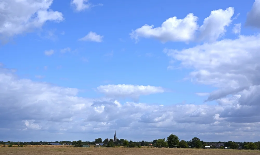 Weite Landschaft mit dem kleinen Dorf Wemb am Horizont unter blauem Himmel mit großen Wolken