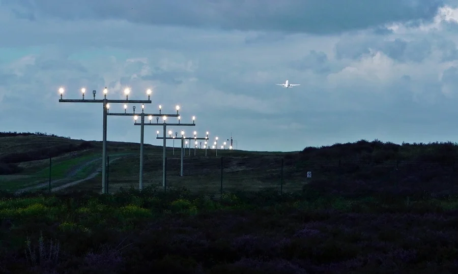 Startendes Flugzeug über der beleuchteten Anflugbefeuerung des Airport Weeze bei dunklem Wolkenhimmel.