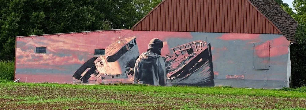 Graffiti von Sébastien Theys auf einer Gebäudewand in Weeze mit Schiffswracks und einer Person im Vordergrund.