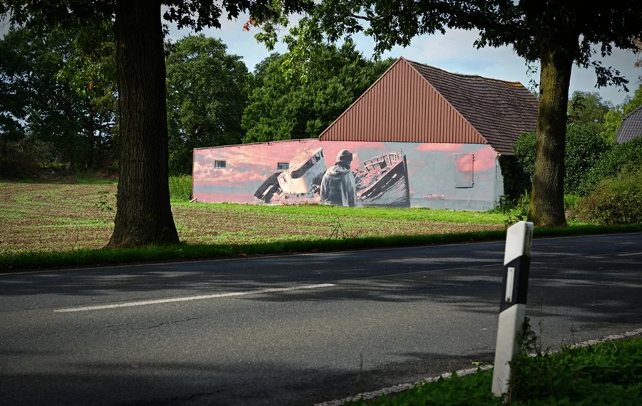 Wandbild an einem Gebäude zwischen zwei großen Bäumen, fotografiert von der gegenüberliegenden Straßenseite