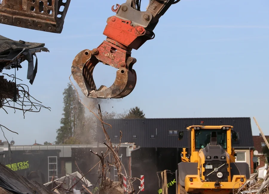 Nahaufnahme einer geöffneten Abbruchzange auf der Baustelle der Johannes-Hauptschule in Weeze. Wasser tropft von der Greifzange, während im Hintergrund eine Baumaschine zwischen den Abrissresten steht.