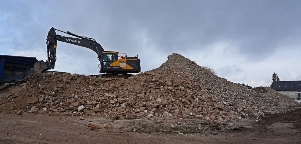 Ein Volvo-Bagger arbeitet auf einem riesigen Berg aus Abbruchmaterial – darüber ziehen dunkle Herbstwolken über die Baustelle in Weeze.