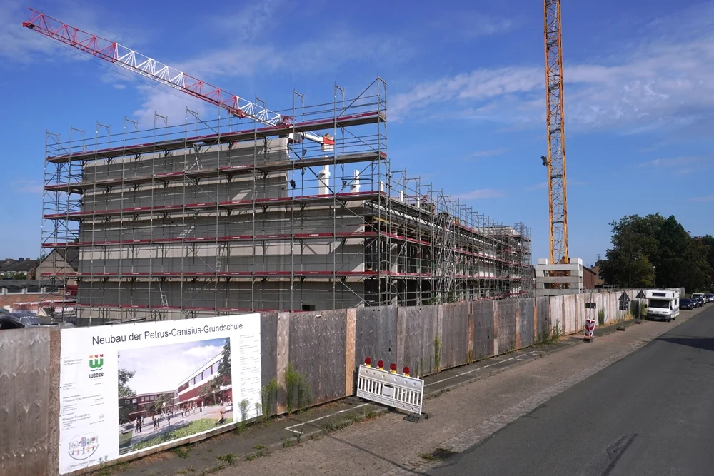 Gesamtansicht der Baustelle der neuen Petrus-Canisius-Grundschule in Weeze entlang der Straße mit Bauzaun, Bautafel, Gerüsten und Baukränen.