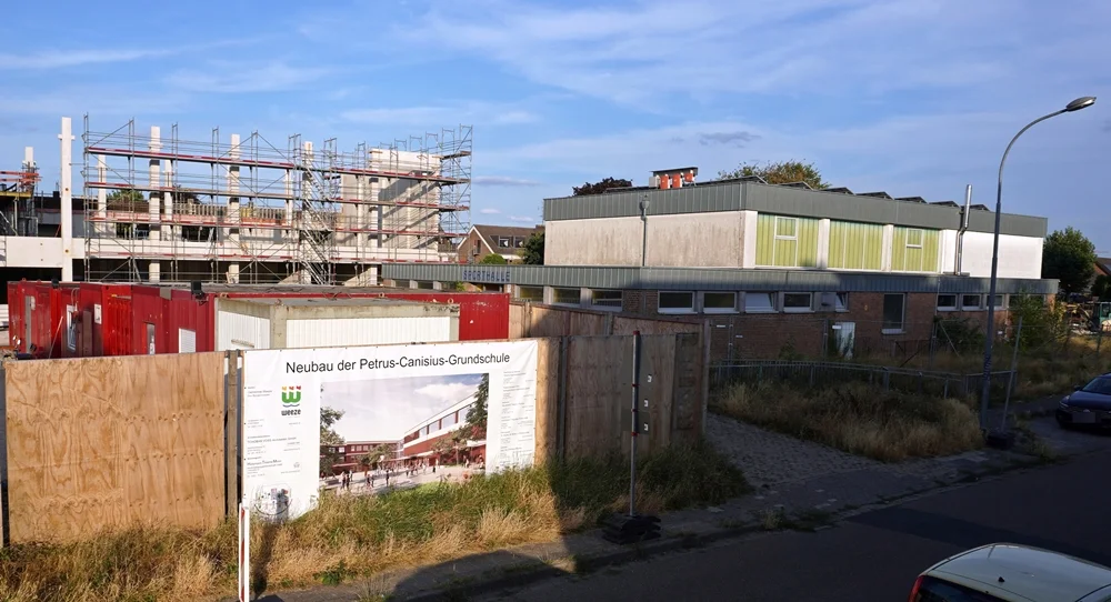 Die alte Sporthalle der Petrus-Canisius-Grundschule in Weeze im Vordergrund. Dahinter ist der Rohbau der neuen Grundschule mit Gerüsten und Baukränen zu sehen.