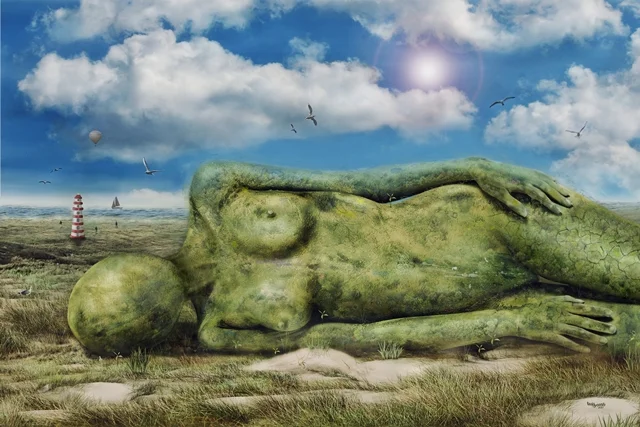 Riesige liegende Frauenfigur aus Stein und Naturmaterialien in einer Küstenlandschaft unter blauem Himmel.