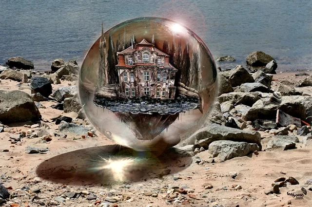 Glaskugel am Strand mit einem kleinen Haus und Bäumen im Inneren. Die Kugel steht zwischen Steinen am Ufer und spiegelt das Licht der Umgebung.