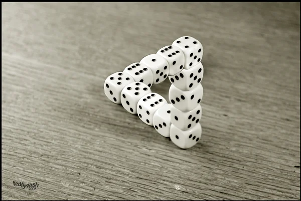 Weiße Spielwürfel bilden auf einer Holzoberfläche ein unmögliches Dreieck. Das Bild spielt mit Perspektive, Geometrie und optischer Täuschung.