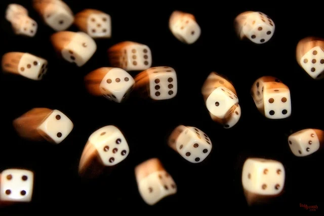Weiße Spielwürfel fliegen vor schwarzem Hintergrund durch die Luft. Die Bewegungsunschärfe vermittelt Dynamik, Zufall und den Moment eines Würfelwurfs.