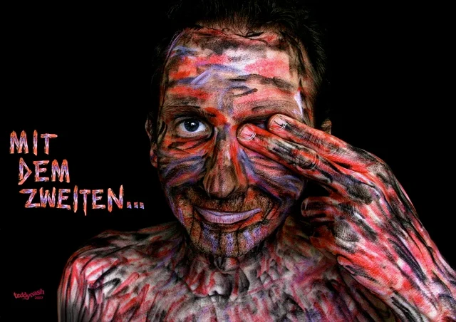 Selbstporträt eines Mannes mit farbig bemaltem Gesicht und Körper vor schwarzem Hintergrund. Mit einem Augenzwinkern verdeckt er ein Auge und blickt direkt in die Kamera - ZDF mit dem zweiten sieht man besser.