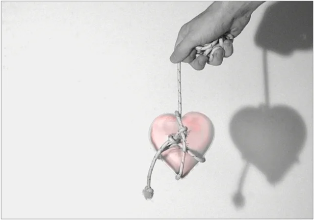 Eine Hand hält ein eingewickeltes Herz an einer Schnur vor hellem Hintergrund. Die reduzierte Szene symbolisiert Liebe, Verlust und emotionale Bindungen.